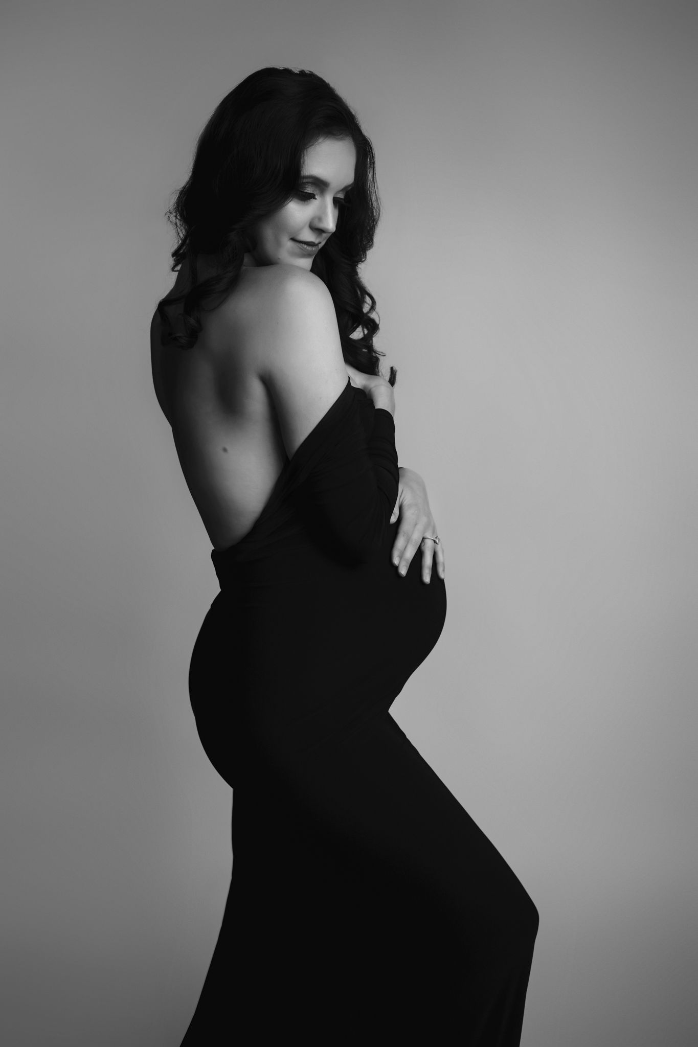 A mother to be stands in a studio in a black maternity gown with her shoulder exposed