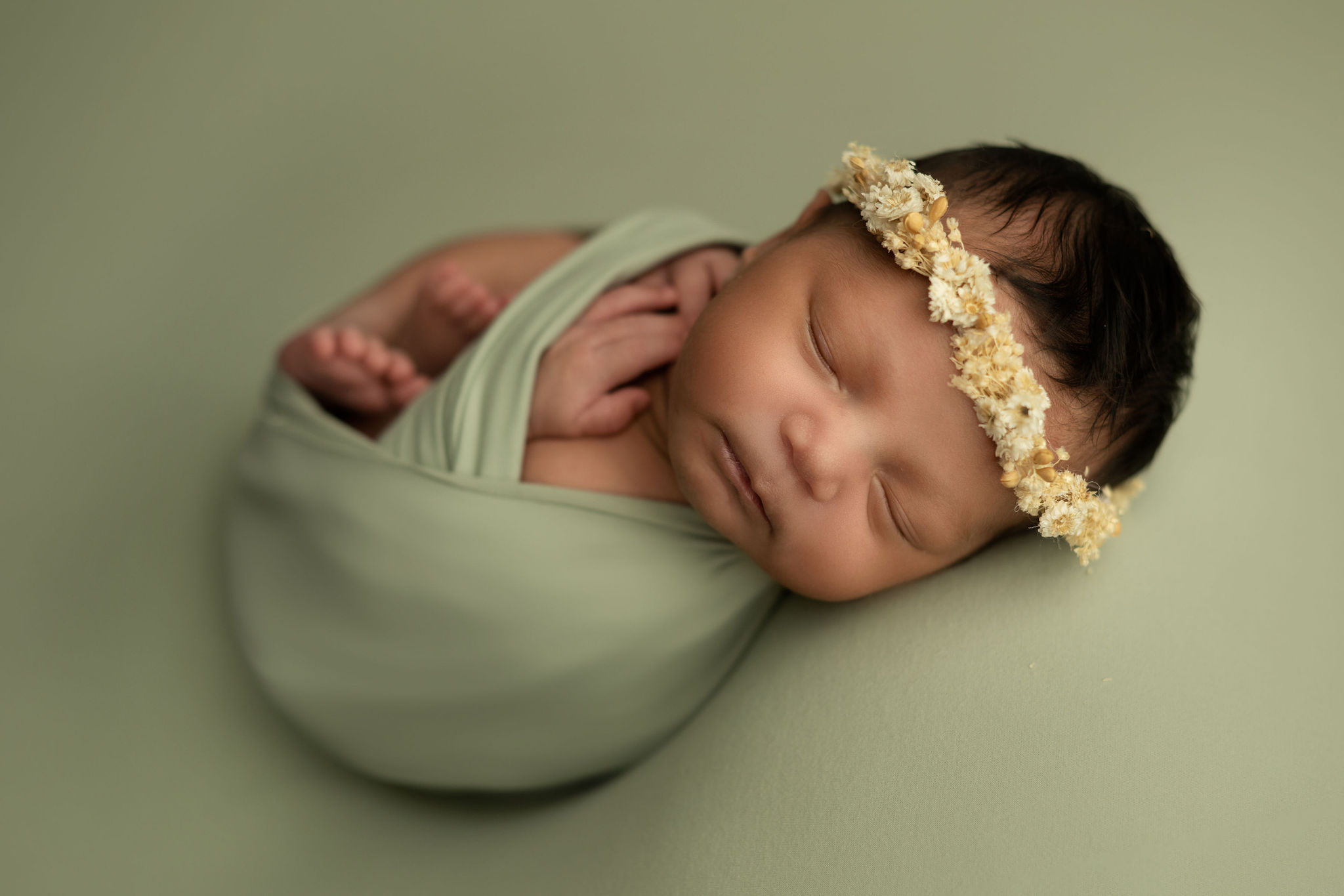 wearing a floral headband, a newborn baby sleeps swaddled in green blanket tampa baby boutique