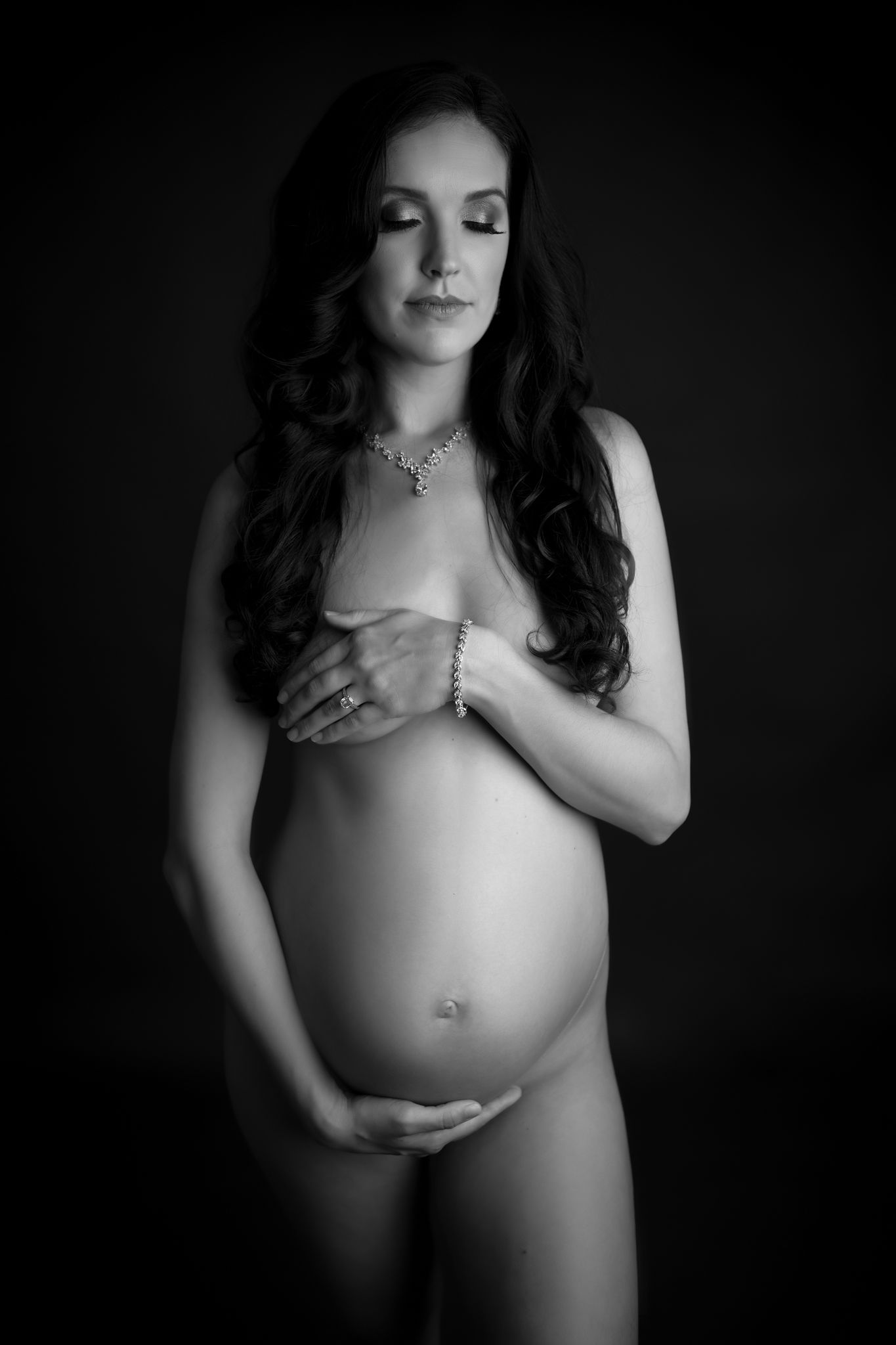 A mom to be stands naked in a studio holding her bump and breasts breath of life birth center
