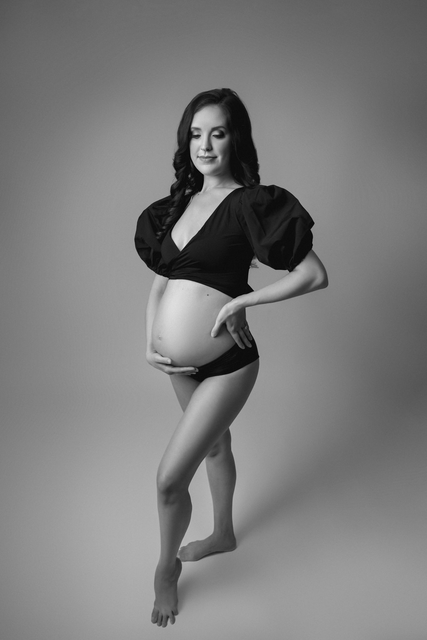 A mother to be in a black top stands in a studio with a hand on her hip and other on her belly