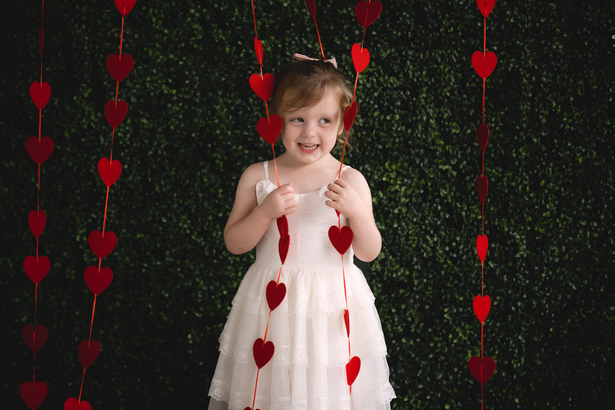 A young girl plays in some hanging heart decorations in front of a greenery wall in a white lace dress tampa toy stores