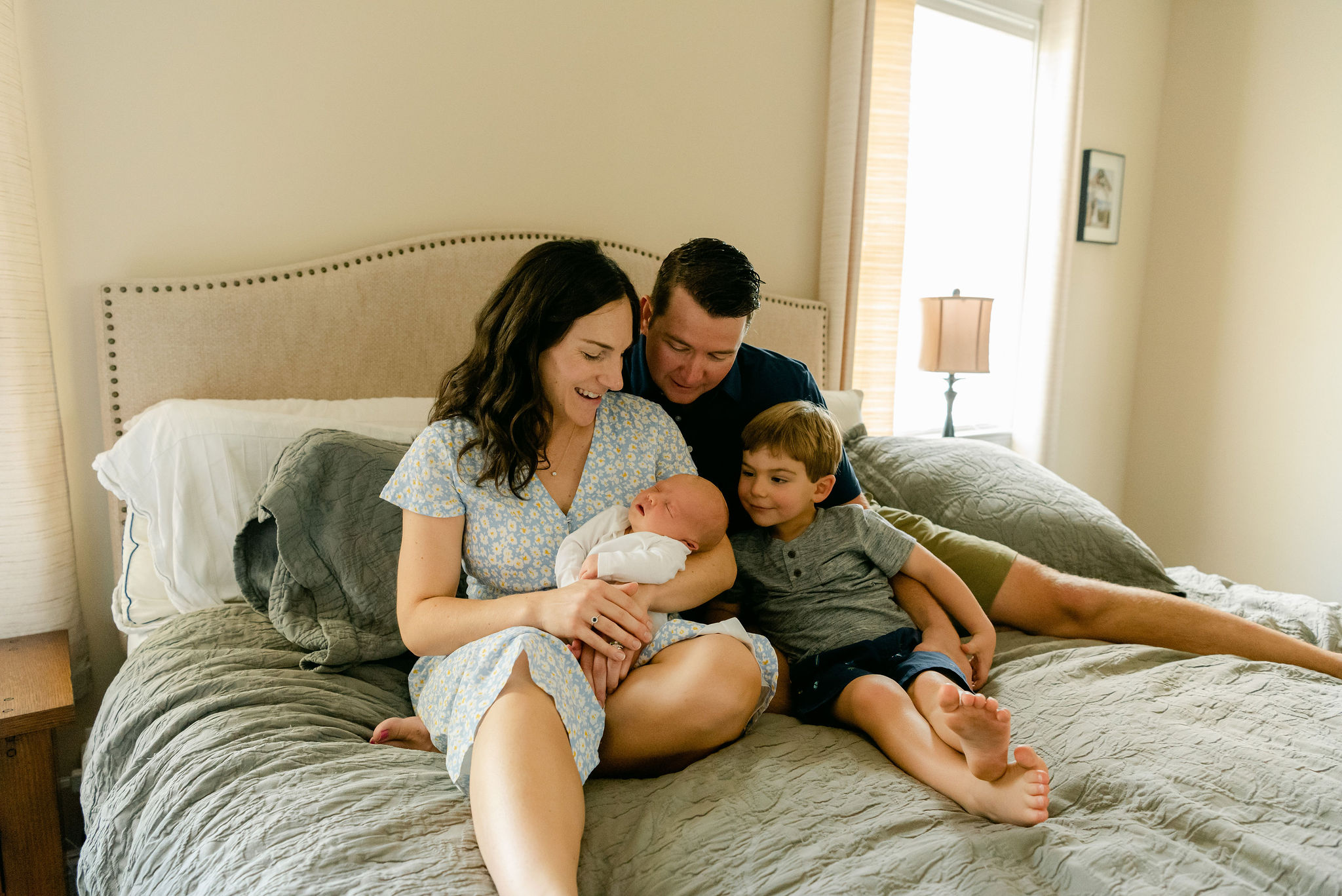 A mother sits on a bed in a blue floral dress holding her newborn baby in her arms while her firstborn son and husband look over her shoulder barefoot birth