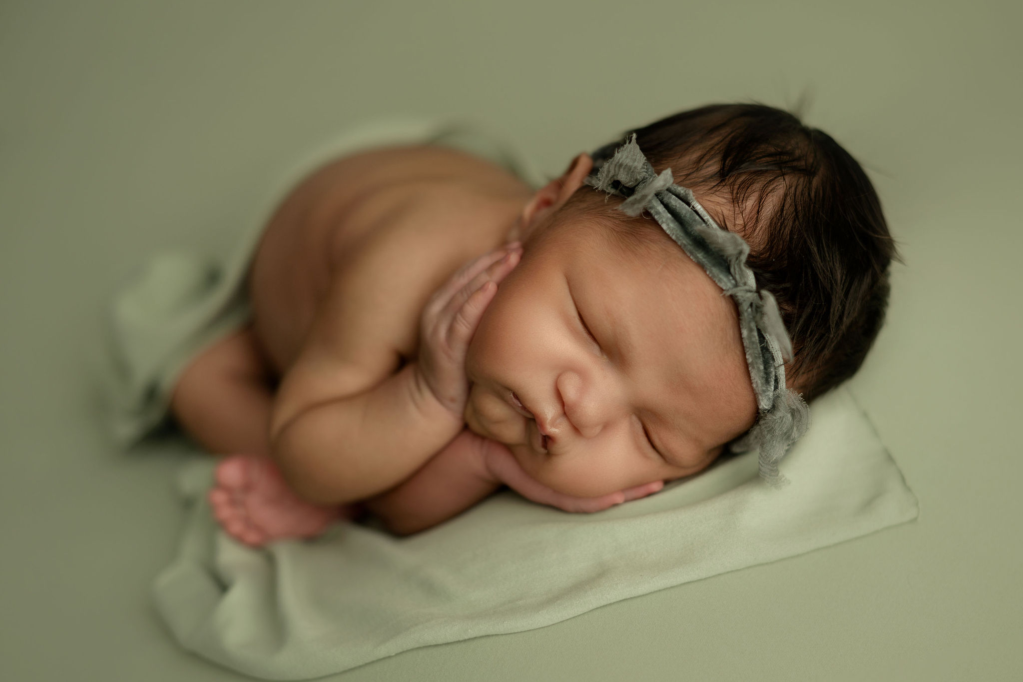 A newborn baby sleeps with hands placed on her cheeks in a green headband