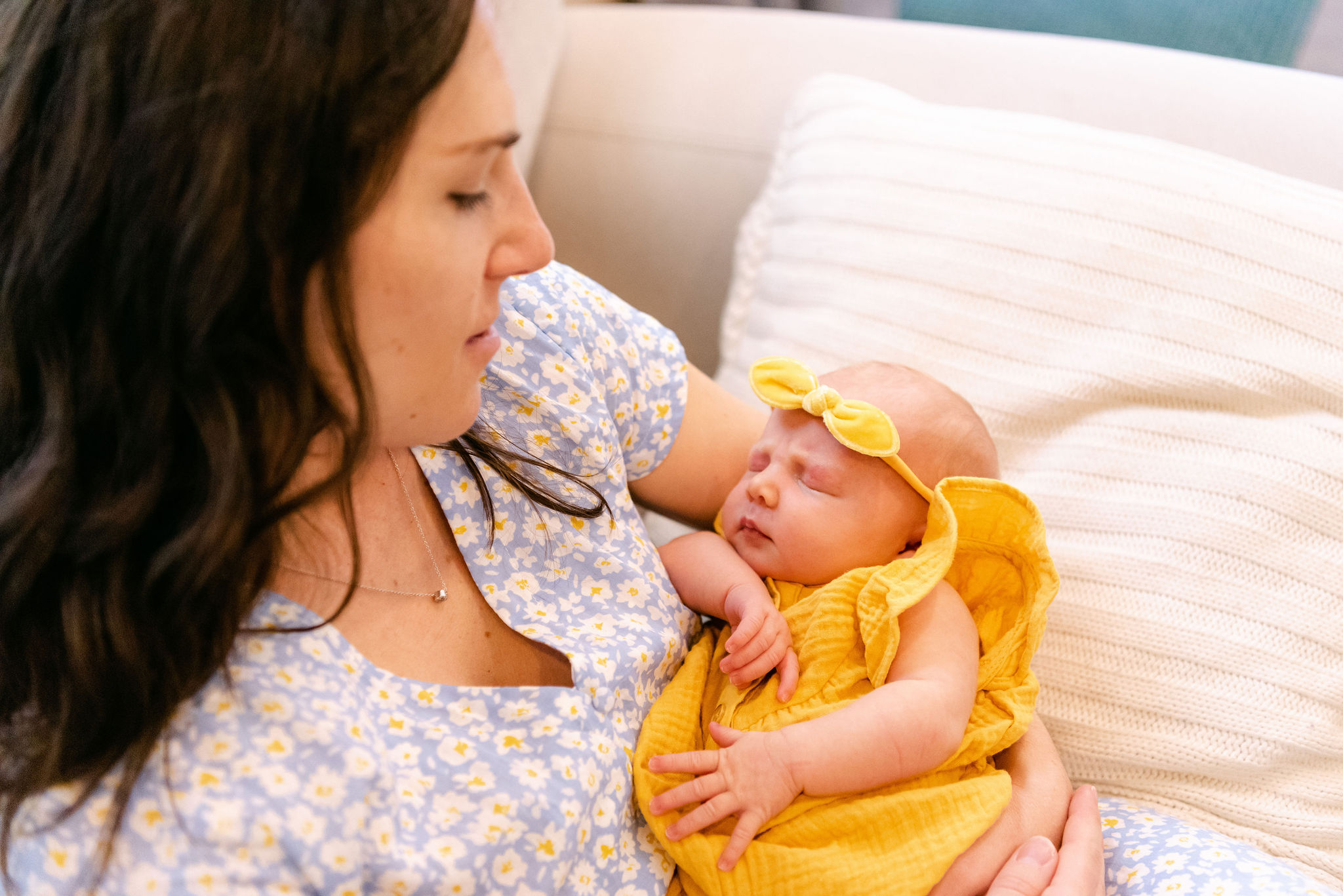A new mother lays on a bed holding her sleeping newborn baby girl in a yellow dress and bow sarasota pediatricians