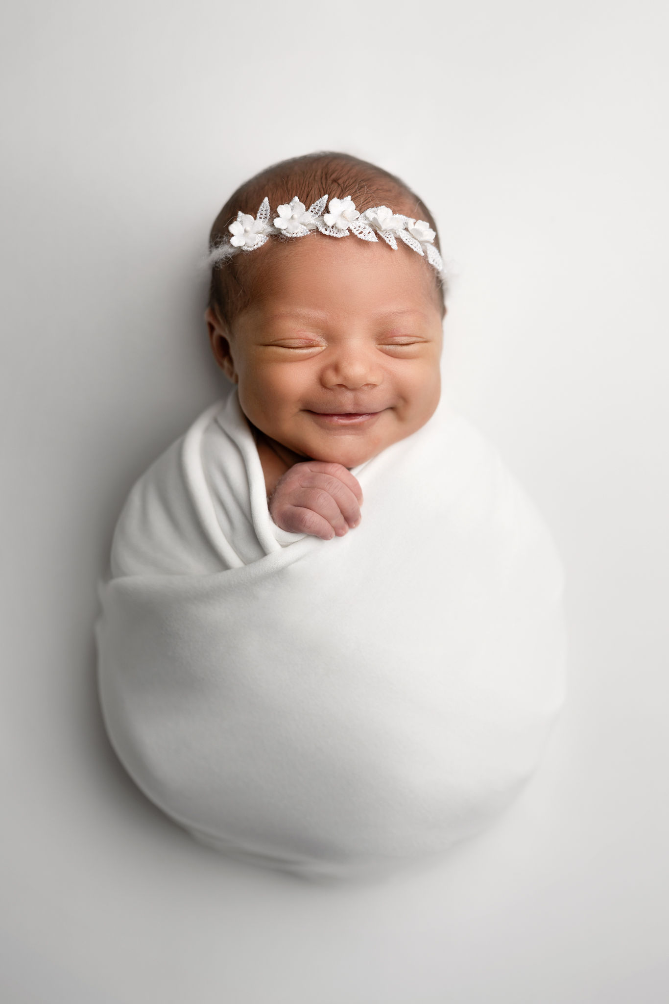 A smiling newborn baby sleeps in a white swaddle
