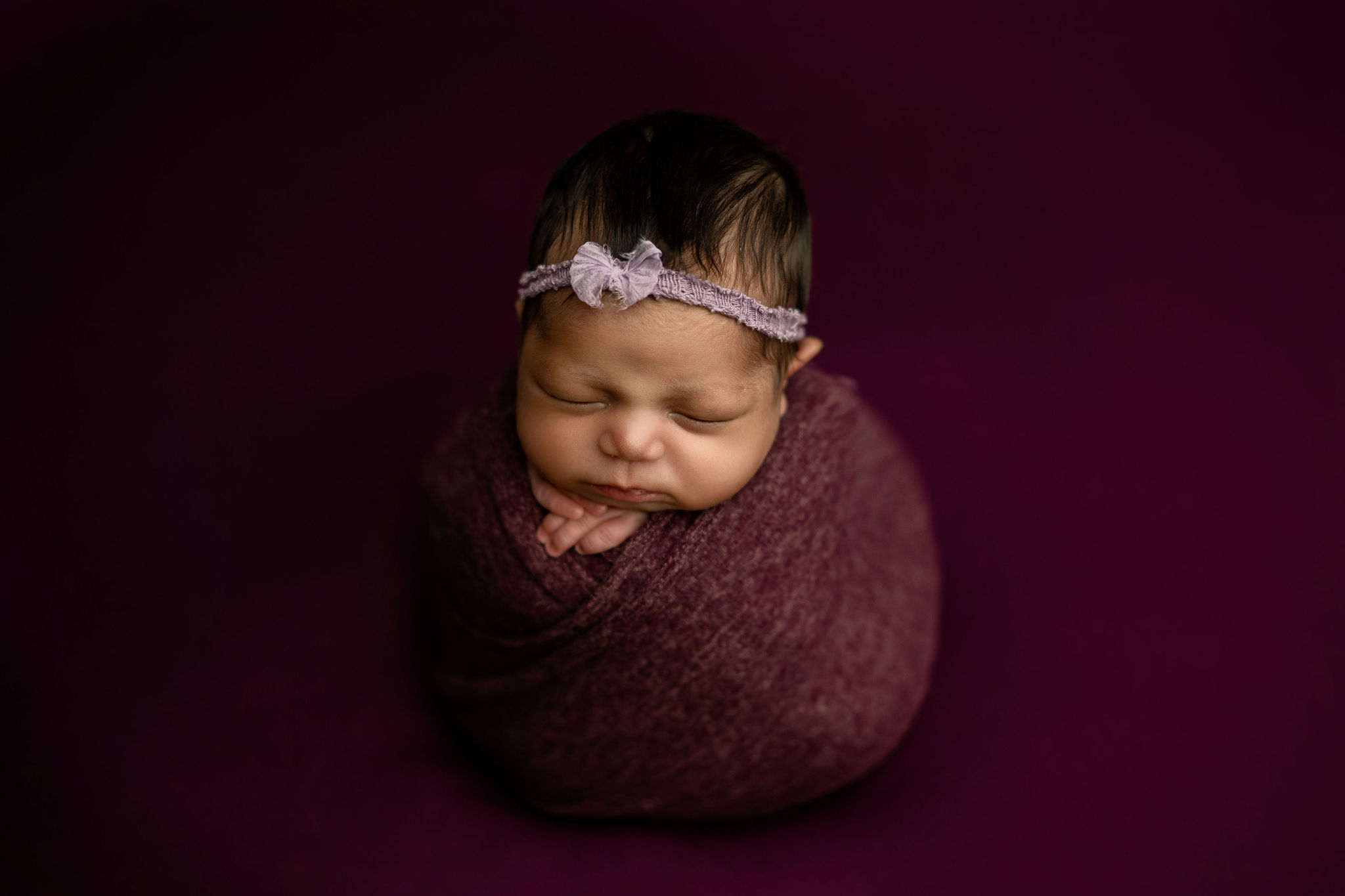 A newborn baby sleeps in a purple swaddle with a light purple headband