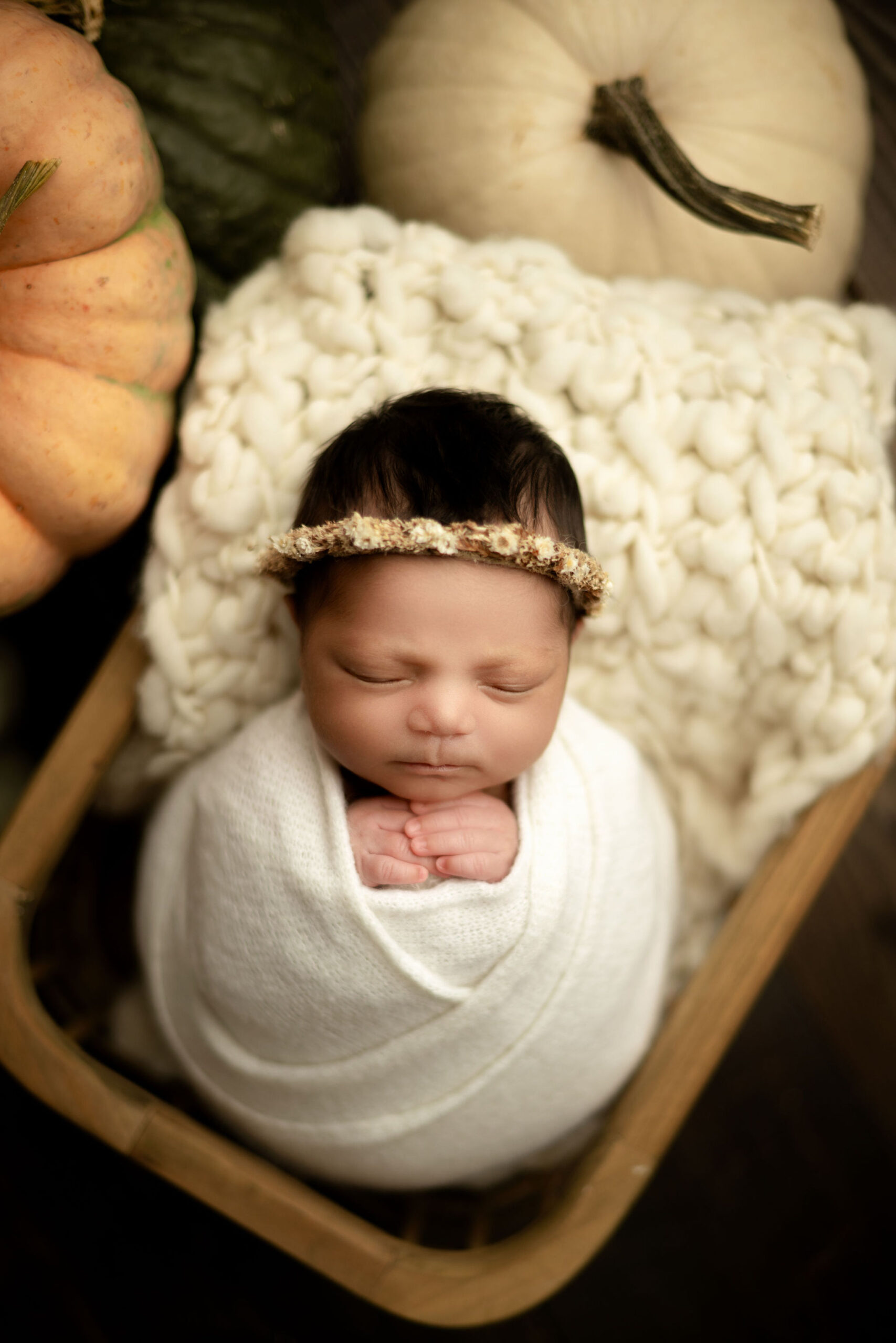 A newborn baby sleeps in a round swaddle with hands poking out surrounded by pumpkins in a wooden basket pottery barn kids tampa