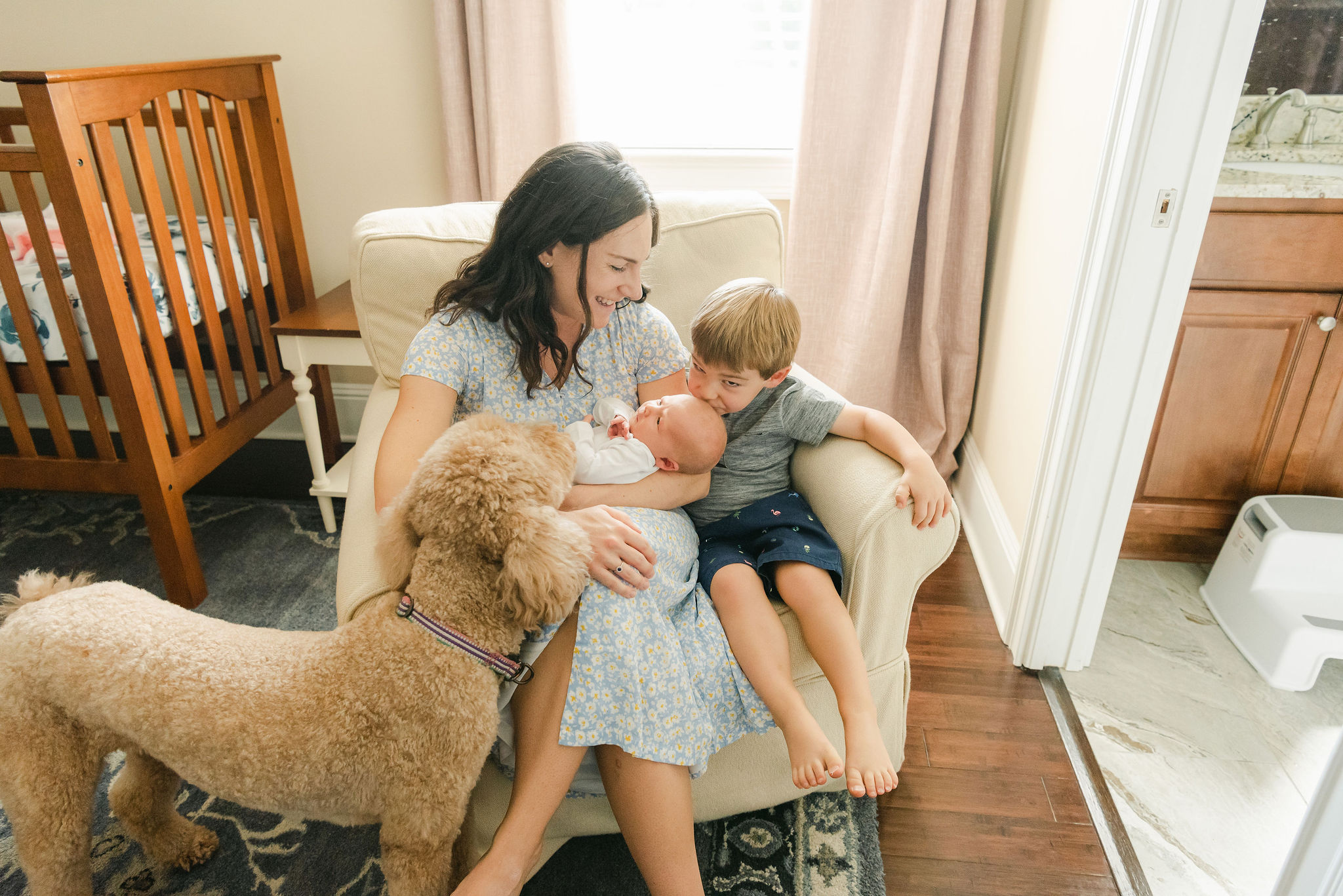 A mother sits on a comfy chair in a nursery with her toddler son and their poodle kissing the newborn baby in her arms